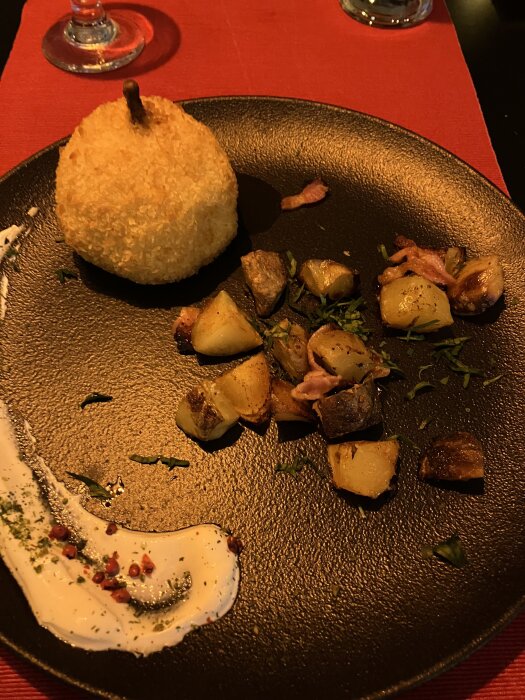 En friterad boll, rostade potatisbitar med örter och skinkbitar, samt en krämig sås med kryddor på en svart tallrik.