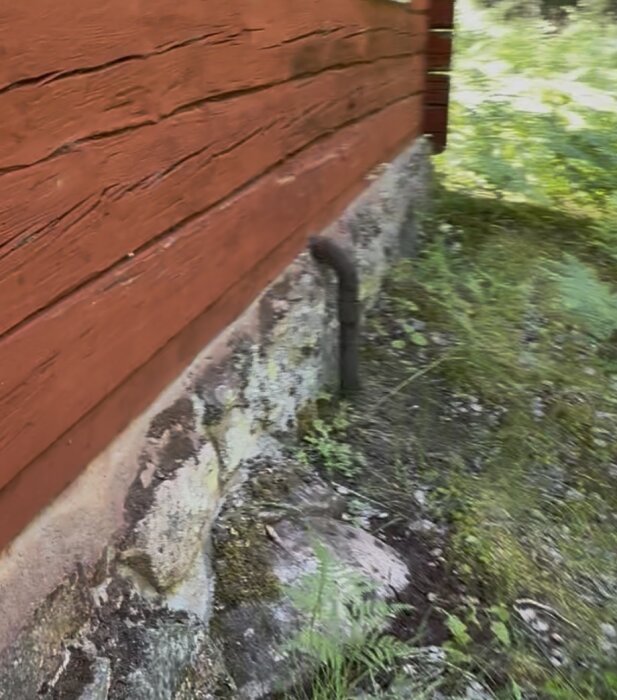 Byggnad med rött träfasad och stenfot, anslutet rör utifrån fundamentet, omgiven av grönska.