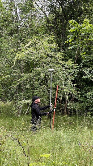 Man i svart kläder använder mätinstrument i grässkog, med ett långt rött och silverfärgat rör för att mäta och lokalisera markpunkter.