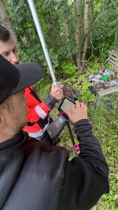 Två personer mäter med utrustning utomhus i ett skogsområde, den ena håller en stång och den andra använder en GPS-enhet.