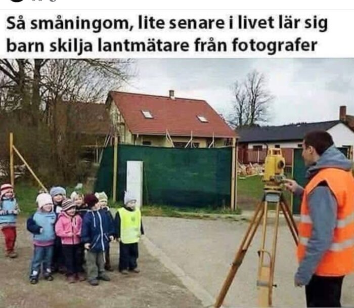 En lantmätare i orange väst mäter med utrustning på stativ medan en grupp barn tittar på som om de poserar för ett foto.