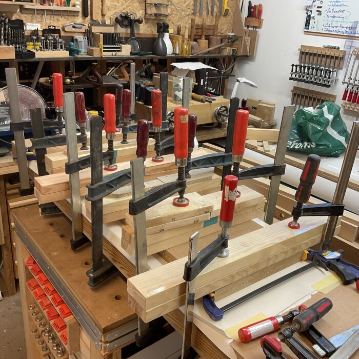 Flera tvingar håller ihop träbitar och MDF i en verkstad, omgivna av verktyg och material.