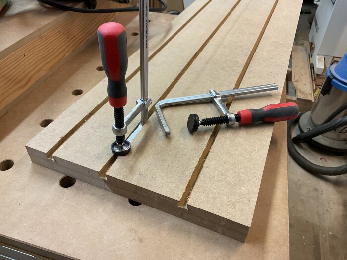 Justerbara klämmor i ett spår på en träbit, omgivna av verkstadsutrustning.