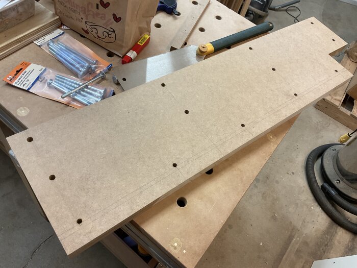 Arbetsbänk med MDF-skivor, förpackningar med vagnsbultar, en såg och limtub. Del av projekt för att skapa högt anhåll till justersåg.