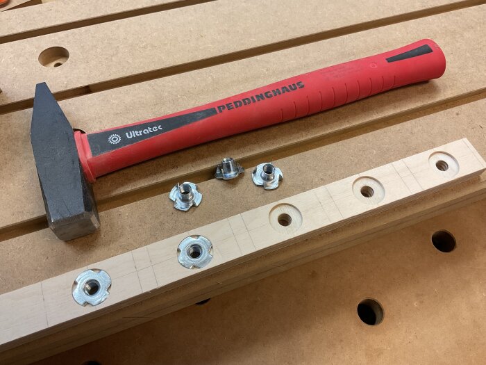 Hammare med rött handtag bredvid en trälist med förborrade hål och islagsmuttrar på en arbetsbänk.