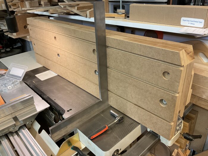 En hög MDF-anhållsdel med spår för vagnsbultar monterad på arbetsbänk, omgiven av verktyg och material i en verkstad.