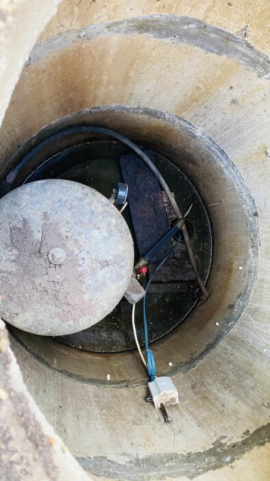 En betongbrunn med kablar och rör inuti, med ett stort lock och vatten vid botten.