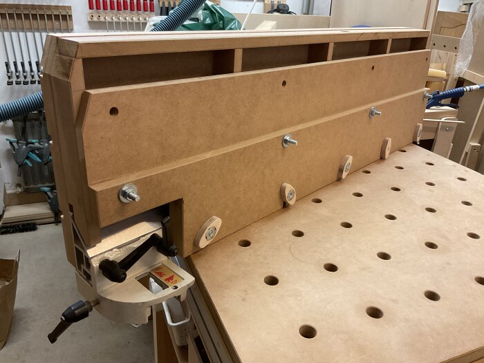 Högt anhåll på justersåg av MDF med plywoodfästen och vagnsbultar på ett perforerat arbetsbord i verkstadsmiljö.