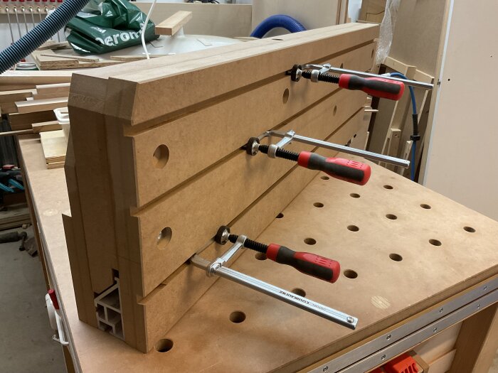 Högt MFD-anhåll på arbetsbänk, fixerat med klämmor. Två halvor sammanfogade och förborrade för montering. Verktyg och träskivor i bakgrunden.