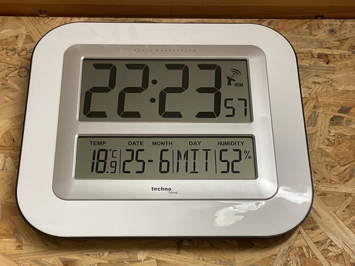Digital klocka med temperatur och luftfuktighet. Tid 22:23, temperatur 18,9°C, datum 25 juni, luftfuktighet 52%. Placeras på trämönstrat underlag.