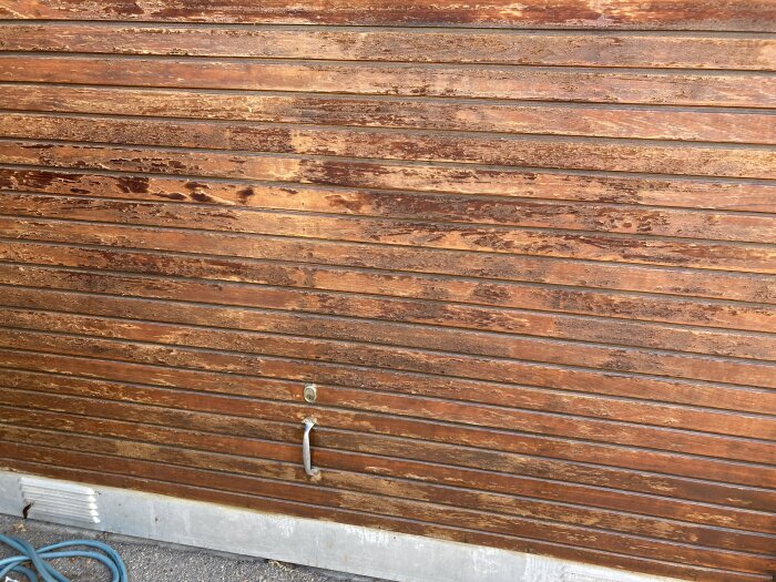 Garageport i trä med synligt slitage och flagnande yta. En slang ligger på marken framför porten.