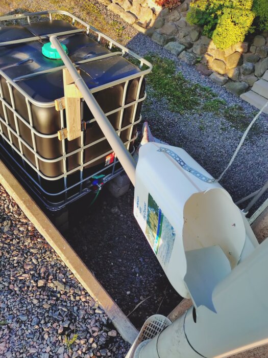 En IBC-tank med improviserad vattenuppsamlingslösning; rörsystem leder vatten ner i tanken från hängränna, omgiven av grus och en stenmur.