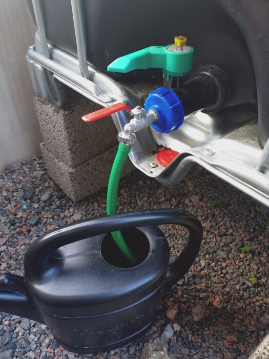 IBC-tank med kran kopplad till en grön slang som fyller en svart vattenkanna. Tankens kran och anslutningar visas tydligt.