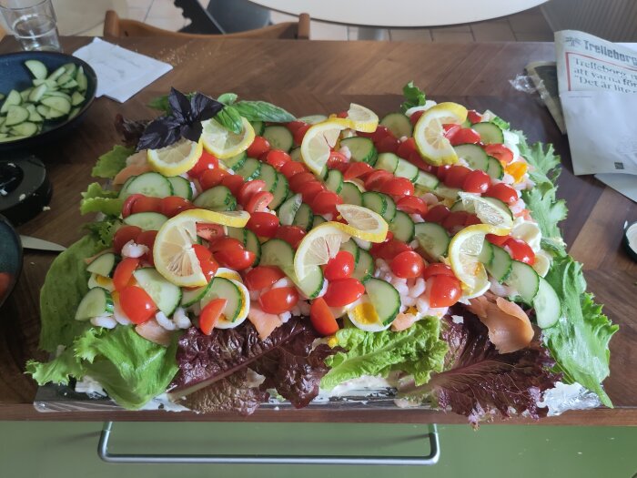 Smörgåstårta dekorerad med sallad, gurka, citron, körsbärstomater, och räkor på ett träbord, omgiven av skålar och papper.