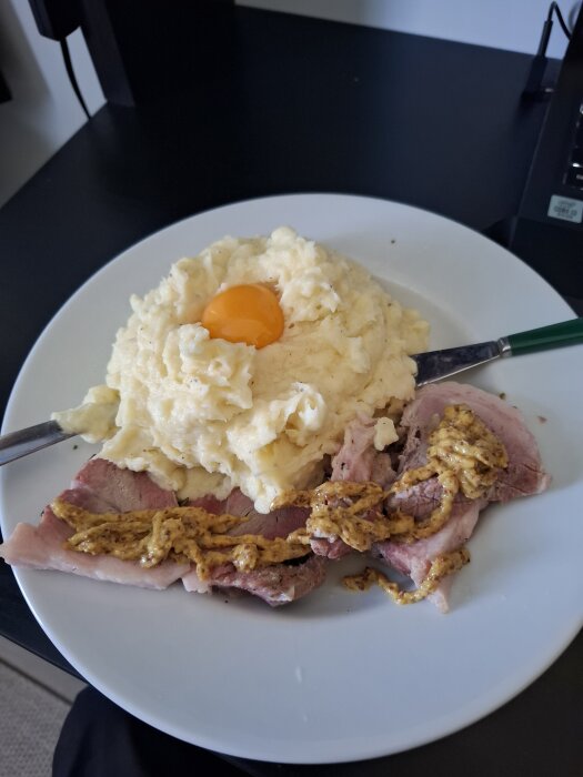 Tallrik med potatismos toppat med ett ägg, serverat med fläsklägg och senap, på ett en svart bordsyta.