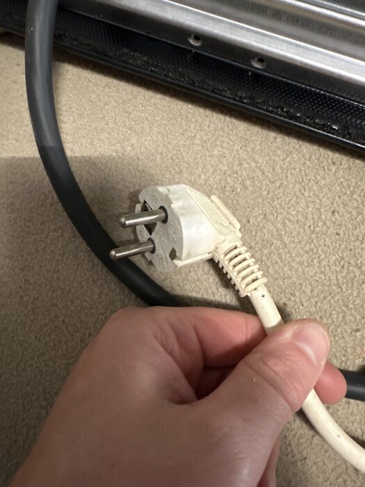 En hand håller i en vit stickpropp med metallstift, uppvikt mot en slät yta. En svart sladd syns ovanför.