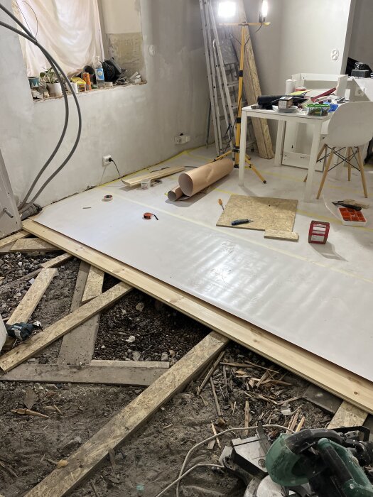 Renovering av rum med golvläggning, verktyg på bord och golv, bygglampor och stege synliga.