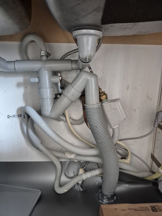 Diskmaskinens avloppsslangar och rör under köksbänk, upphängda med många böjar. Droppskydd och kopplingar synliga.