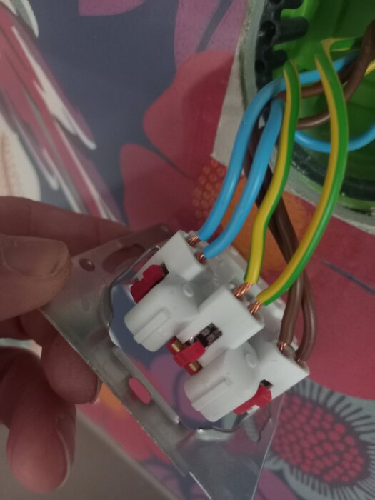 Elektrisk kopplingsdosa med synliga kablar, kopplad i en vägg med färgglad tapet. En hand håller i dosan.