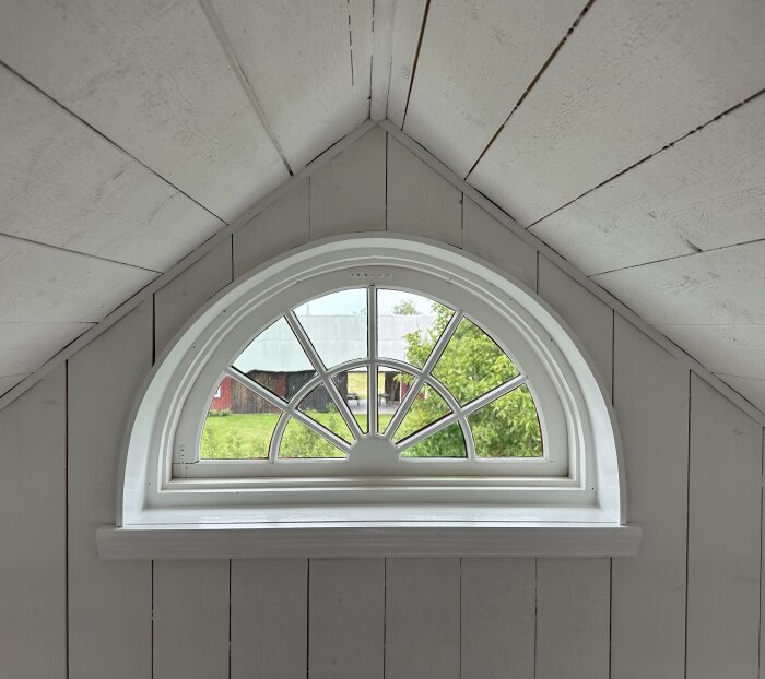 Halvmåneformat fönster i vitmålad trävägg, med utsikt över en gård och grönt landskap utanför.