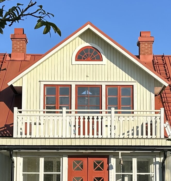 Fasad på ett hus med gul panel och rött tak. En vit balkong och fönster med röda karmar syns tydligt.