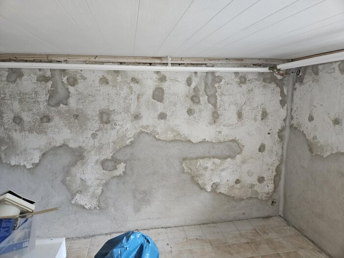 Ojämn källarvägg med fläckar av cement och putsbruk, skarvar och strukturer synliga. Tegelgolv och tak samt röret upptill i bilden.
