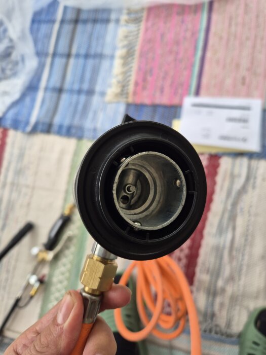 Gasolregulator med orange slang i närbild, hålls i handen. Bakgrunden visar en färgglad matta och oskarpa föremål.