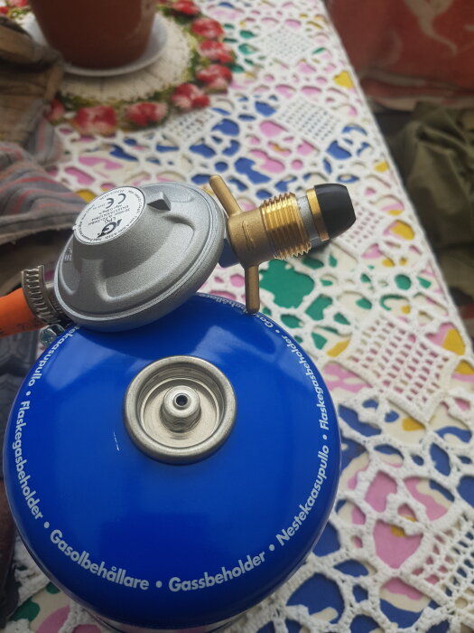 Regulator kopplad till blå gasolflaska på färgglad duk, sök efter adapter för korrekt anslutning.