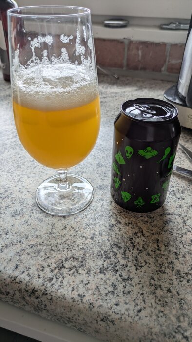 Glas med orange dryck bredvid en svart burk med gröna rymdmotiv, placerade på en köksbänk med tegelvägg i bakgrunden.