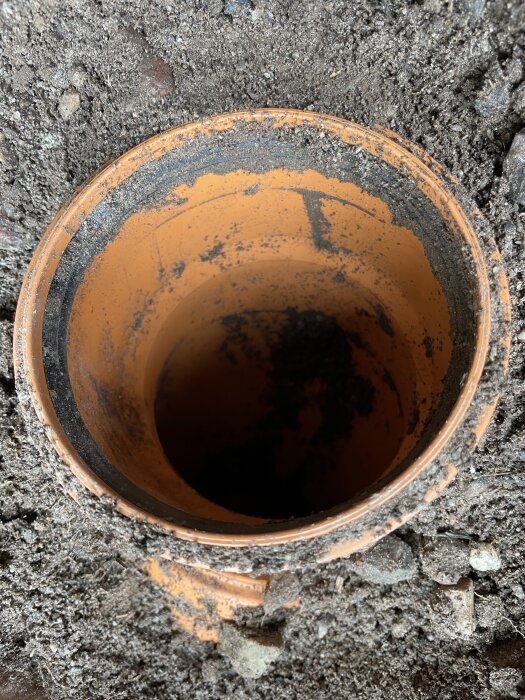 Orange plast- eller lerör i marknivå, omgivet av jord och grus. Rörets insida synlig, delvis fylld med jord.