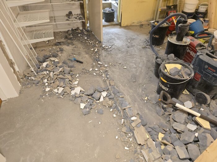 Rester av lättbetong och verktyg på ett dammigt golv, med hinkar och en dammsugare i bakgrunden under renovering.