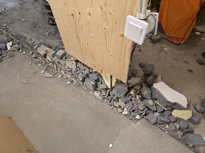 Rester av rivningsarbete, lättbetong och murbruk synliga längs en ränna under en plywoodvägg med eluttag monterat.