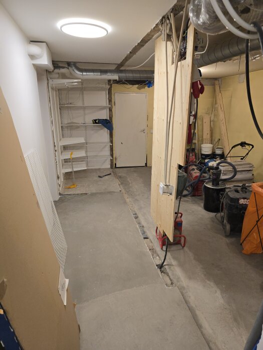 Källare under renovering; en påbörjad vägg, rör, hyllor och rengöringsutrustning syns. Snett tak och konstruerad innervägg.