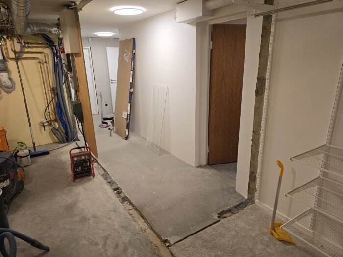 Hall med synliga rör och en halvvägg under renovering; en ränna i golvet där en vägg fanns. Dörr leder till annat rum, verktyg och redskap synliga.