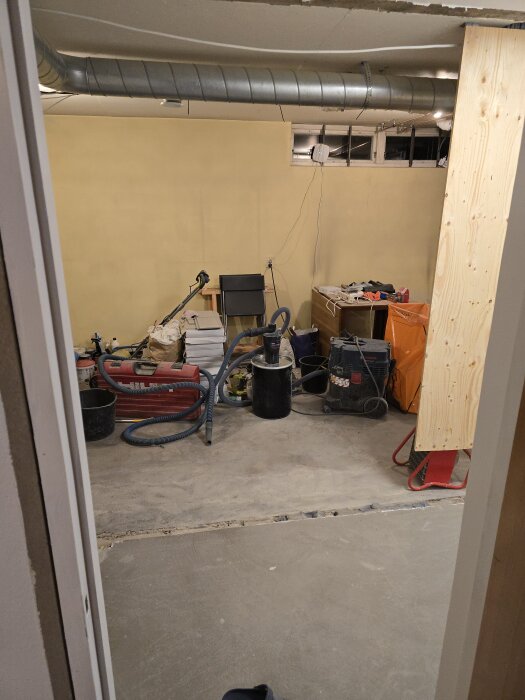 Renoveringsprojekt med verktyg och utrustning i ett rum med gul vägg och ventilationsrör, plywoodskiva, dammsugare och öppna lådor runtom.