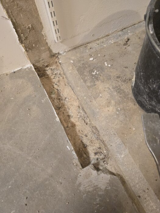Ränna i betonggolv där en innervägg tagits bort, omgiven av smutsigt underlag med murbruk, en vägg hörn och en svart hink bredvid.