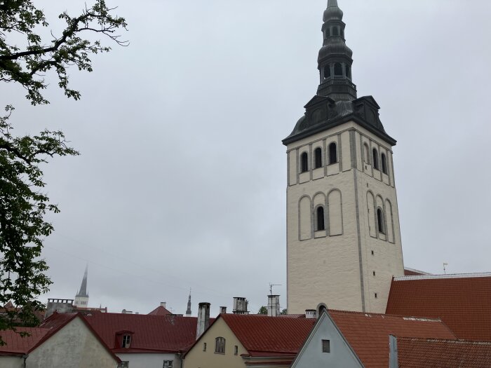 Kyrktorn i stadsmiljö med röd taknock i förgrunden och träd vars grenar når in från vänster sida, mot en molnig himmel.