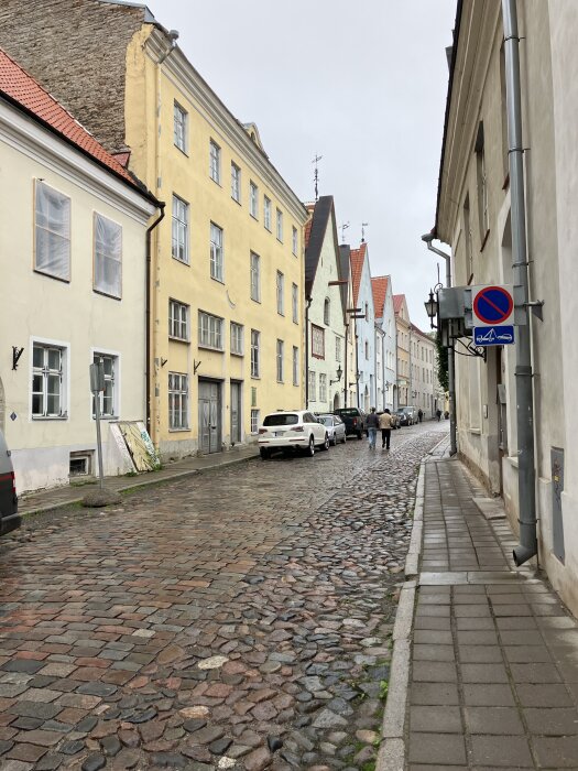 Kullerstenbelagd gata i gammal stad med färgglada hus och bilar parkerade längs trottoaren, två personer promenerar i fjärran.