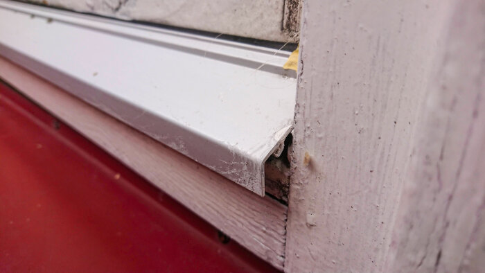 Aluminiumprofil på altandörr med skadad kant och synlig trästruktur. Tätningslist av gummi finns under profilen.