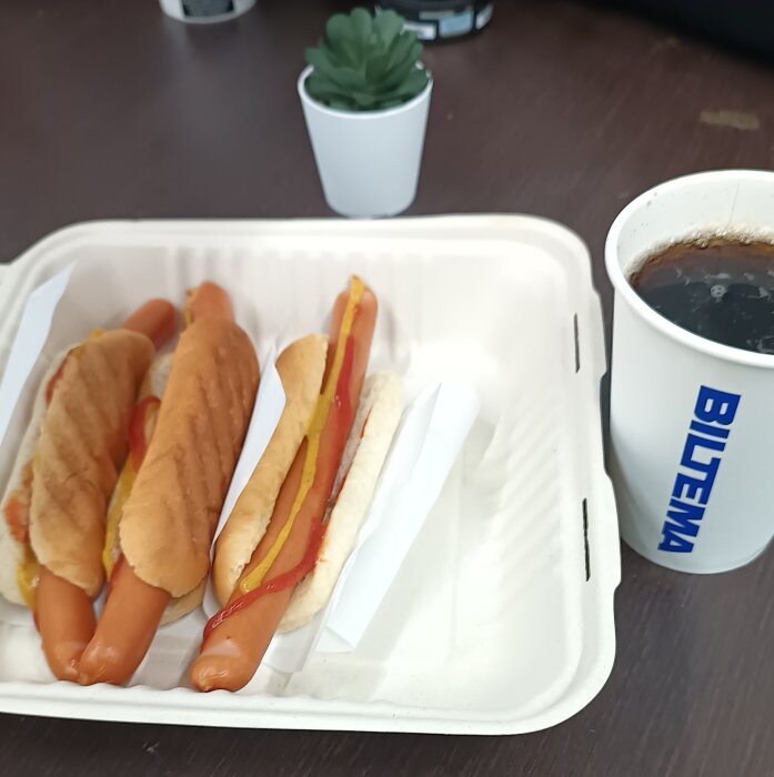 Tre varmkorvar i bröd med ketchup och senap i en kartong, en pappersmugg med dryck och en liten grön växt på ett bord.