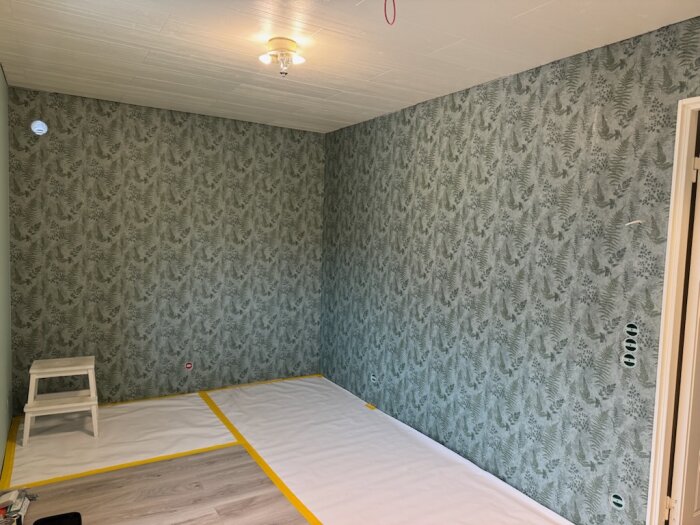 Vägg rumsdesign med bladmönstrad tapet, golv täckt med skyddspapper, stege mot väggen, förberett för inredning med ny tapet.