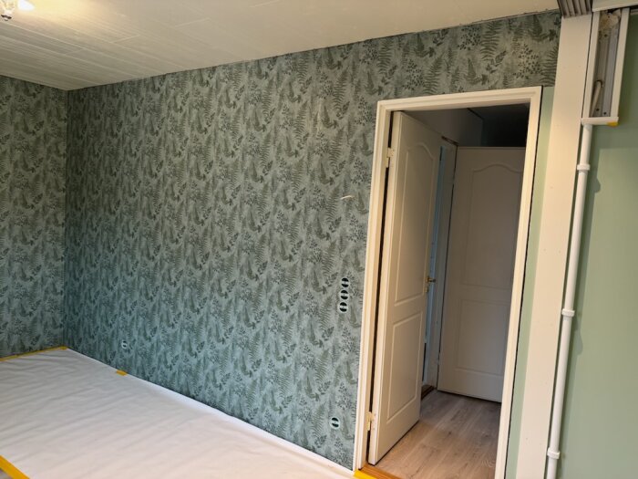 Grönt tapetserat rum med mönster av växter, öppen dörr och synliga rör längs väggen.