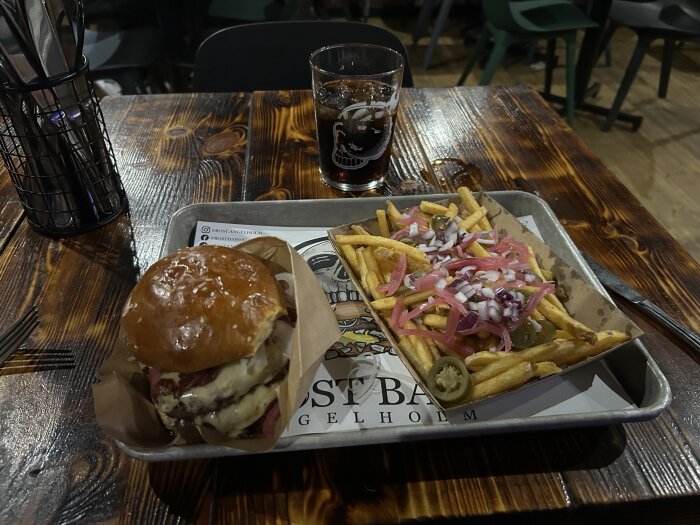 Hamburgare och pommes frites med lök, jalapeños och picklad rödlök på en bricka, serverat med ett glas dryck på ett träbord.