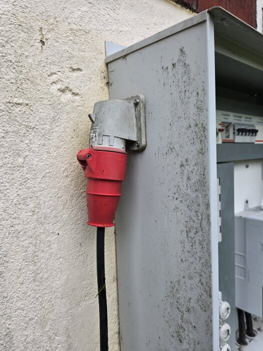 Röd elektrisk kontakt ansluten till ett elskåp på en utomhusvägg, omgiven av smuts och slitage.