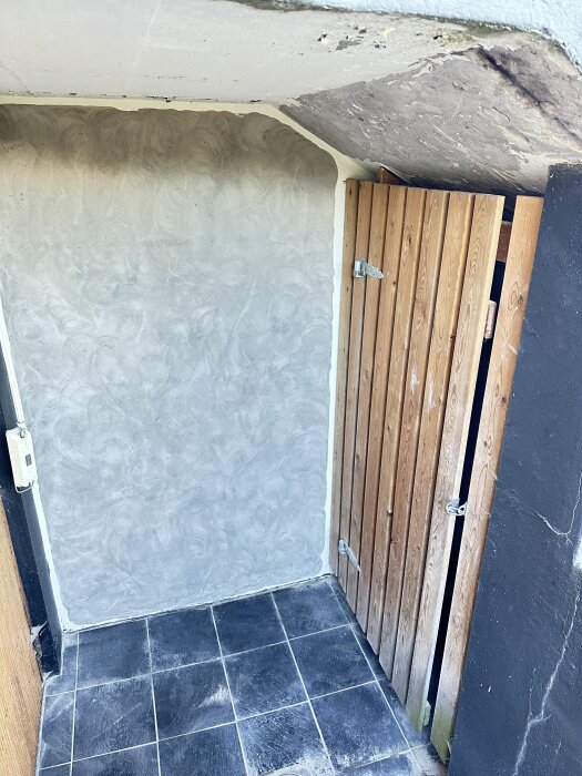Renoverat hörn av ett rum med träpanel och ljusgrått putsat tak och vägg, svart golv av kvadratiska plattor.