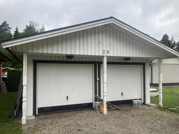 Vit garage med två portar, nummer 29, utan dekorativa detaljer. Finns målburkar och redskap framför i grusuppfart.