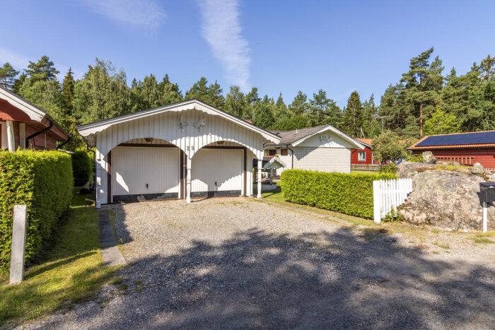 Garage med borttagen utsmyckning, omgiven av gröna buskar och staket. Soligt väder och skog i bakgrunden.