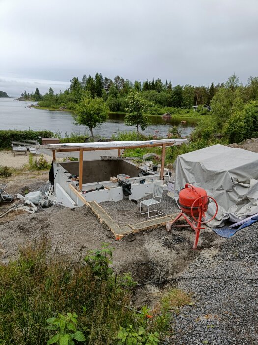Byggarbetsplats vid sjö, förberett område för pooldäck och stödmur med betongblandare och utsikt över träd och vatten.