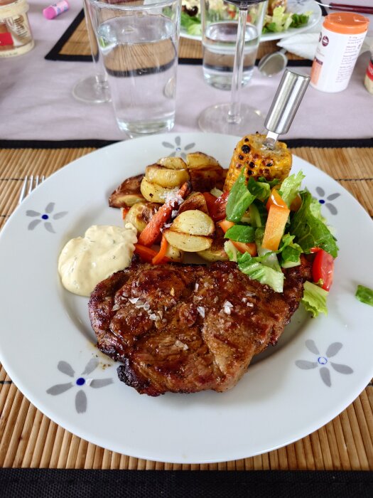 Grillad karré med färskpotatis, ugnsmorötter och majskolv på tallrik, serverad med sallad och sås.