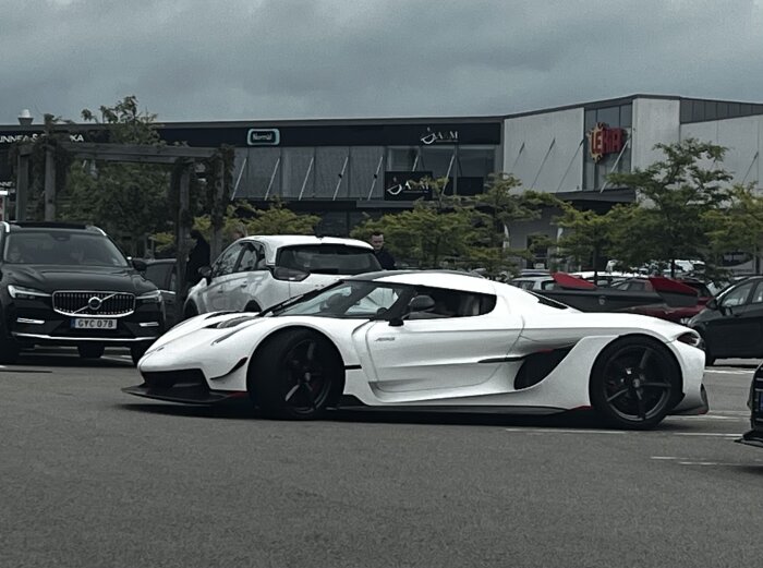 Vit sportbil parkerad på en parkeringsplats, omgiven av andra bilar, med ett köpcentrum i bakgrunden under en molnig himmel.